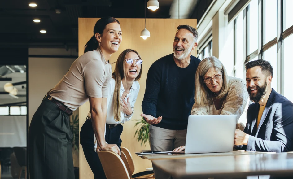 Five colleagues laughing together around a laptop in a bright office
